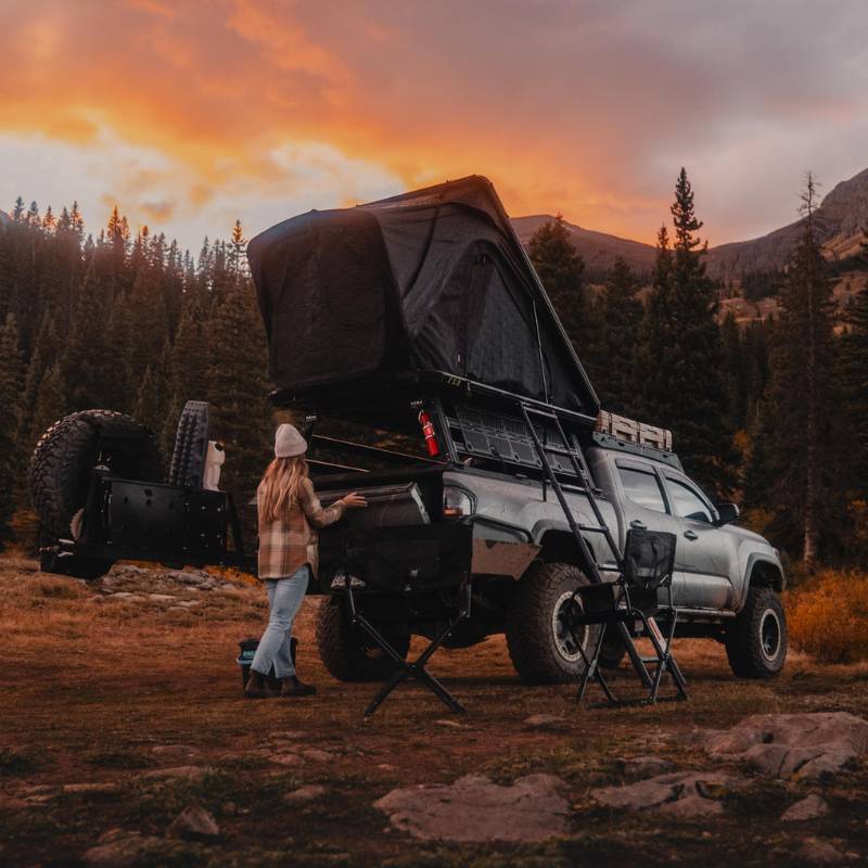 freespirit-recreation-aspen-v2-xl-hard-shell-roof-top-tent-black-open-rear-corner-view-on-toyota-tacoma-with-woman-and-chairs-in-nature