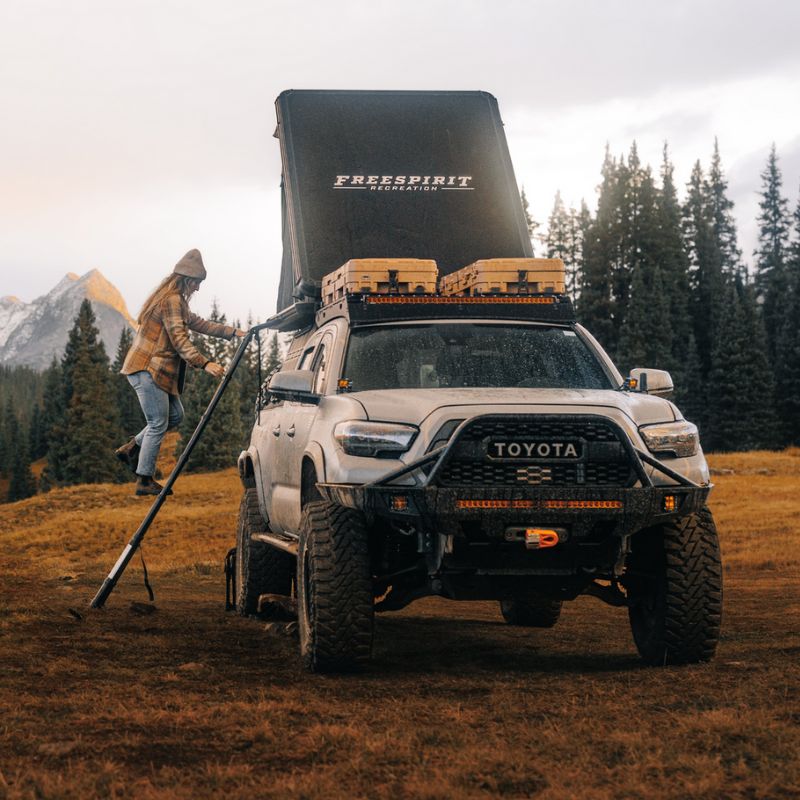 freespirit-recreation-aspen-v2-hard-shell-roof-top-tent-black-open-front-view-on-toyota-tacoma-with-woman-climbing-ladder-in-nature