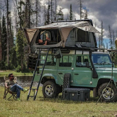 overland-vehicle-systems-bushveld-hard-shell-roof-top-tent-open-front-corner-view-on-land-rover-defender-at-lake-side-with-persons-camping