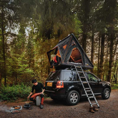 tentbox-cargo-hard-shell-roof-top-tent-black-open-front-corner-view-on-vehicle-with-man-cooking-and-person-inside-tent-in-nature
