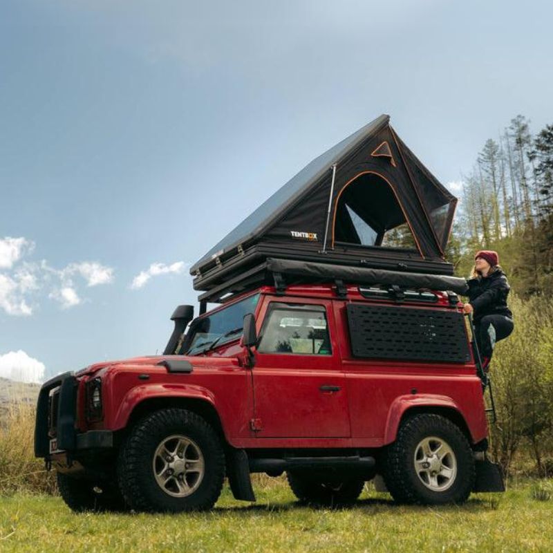 tentbox-cargo-hard-shell-roof-top-tent-black-open-side-view-on-land-rover-defender-with-woman-climbing-ladder-in-nature